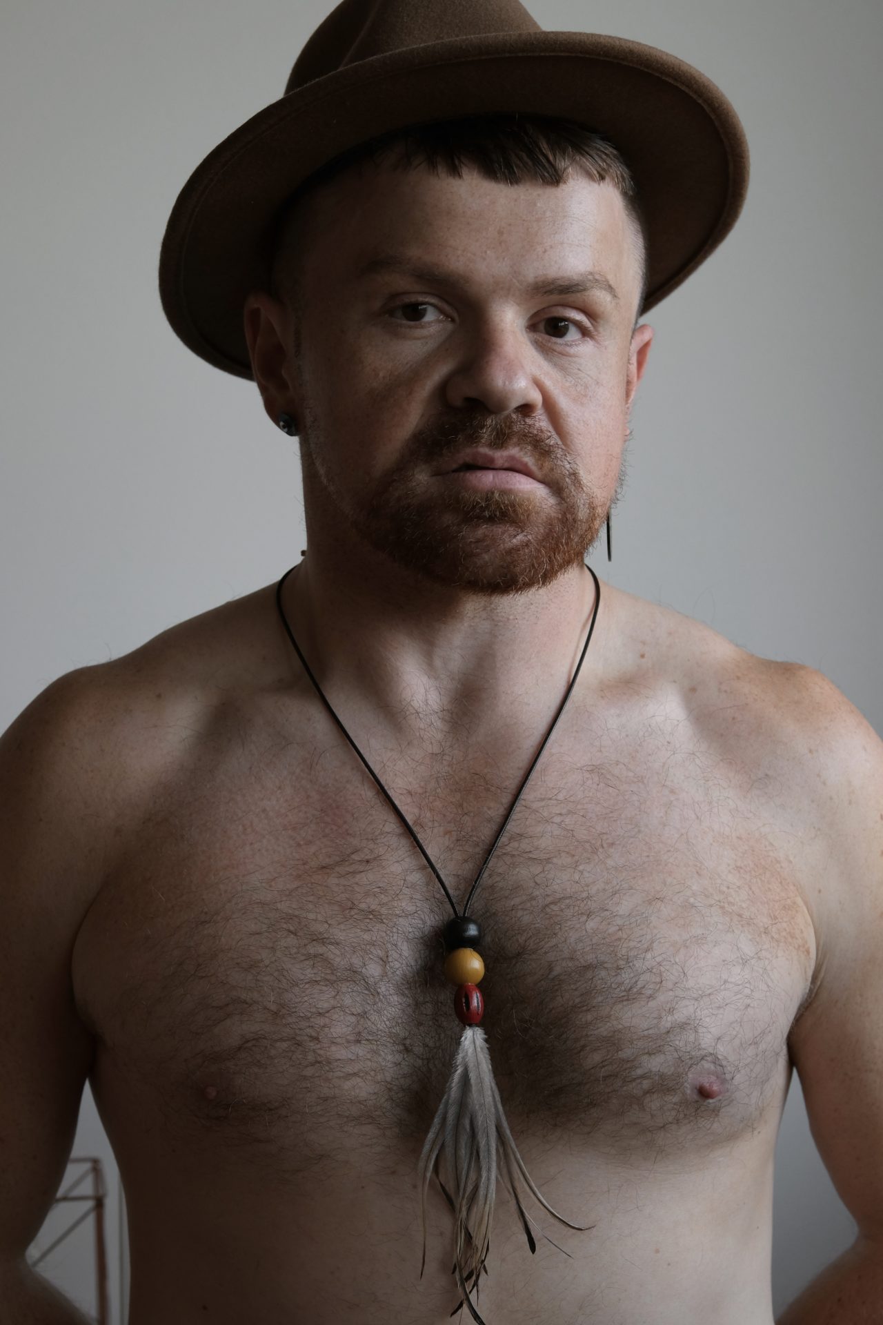 Will Cooper poses for the camera shirtless with a brown hat and an emu feather necklace.