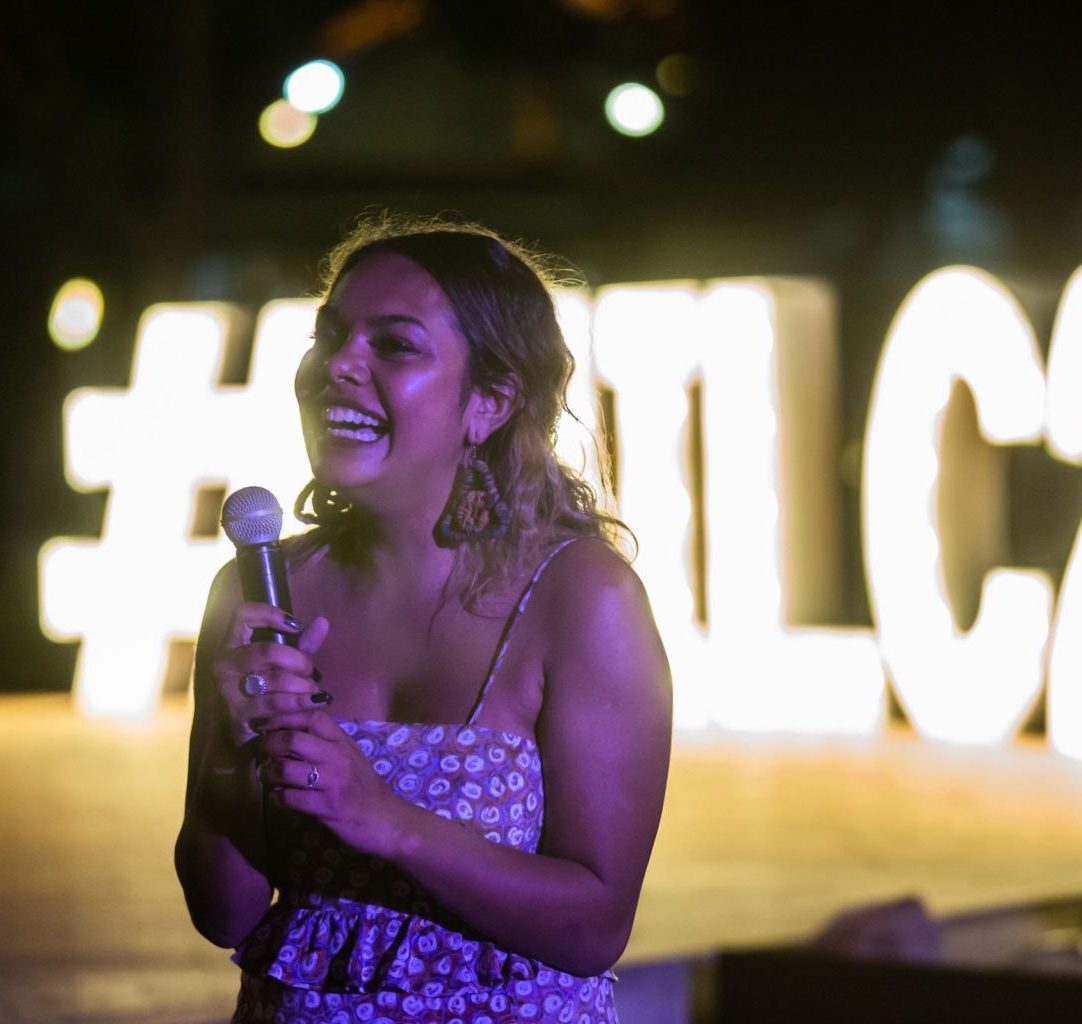 A First Nations person stands with a microphone in front of some neon letters.