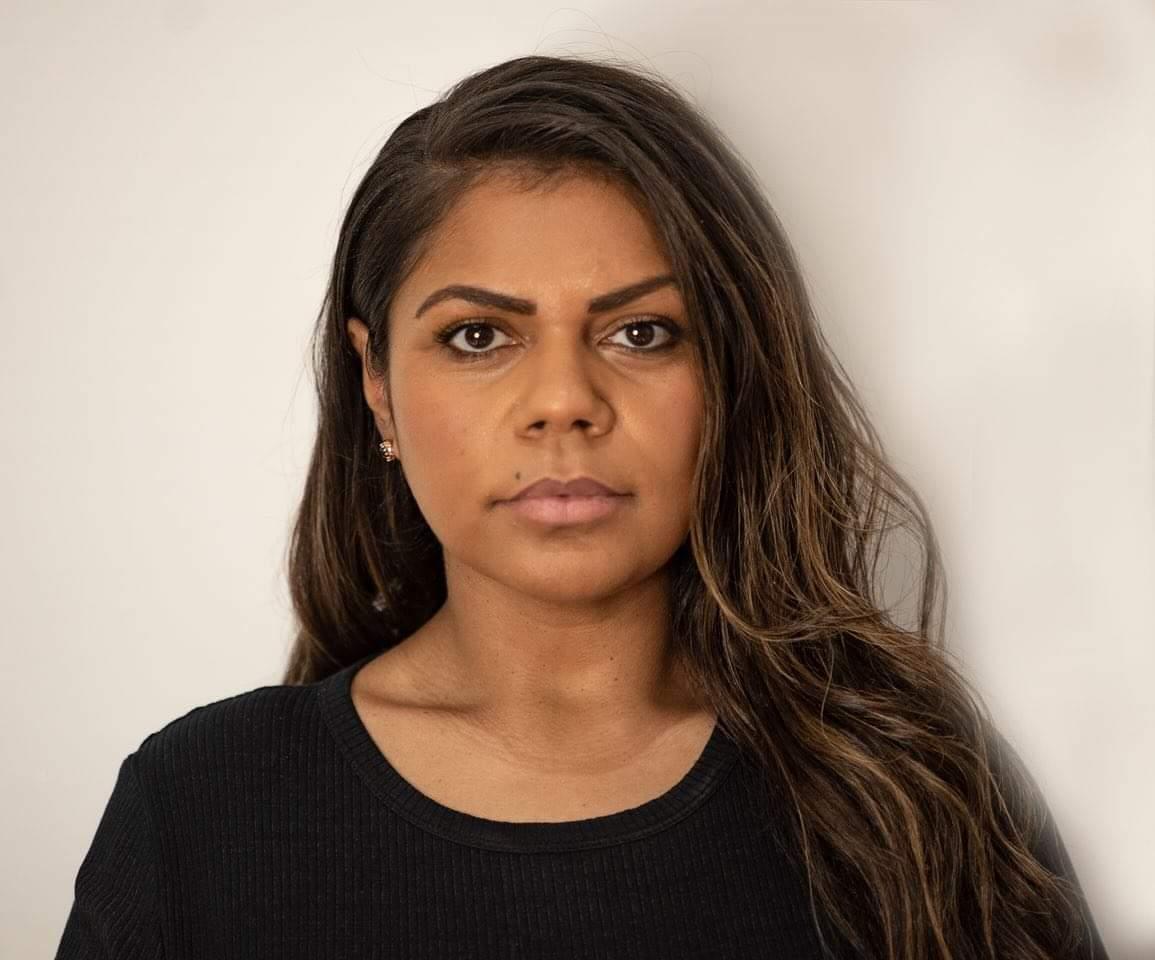 A First Nations person with long hair stands in front of a white wall.