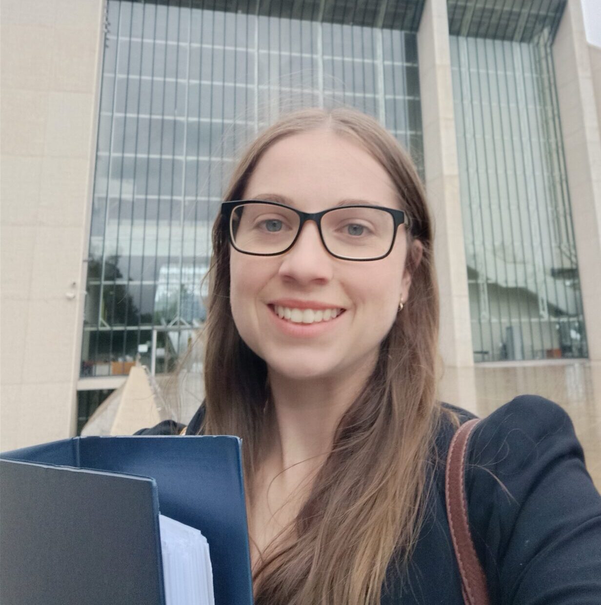 Karina is a person with long brown hair and glasses. They are smiling in the photo and folding a folder.
