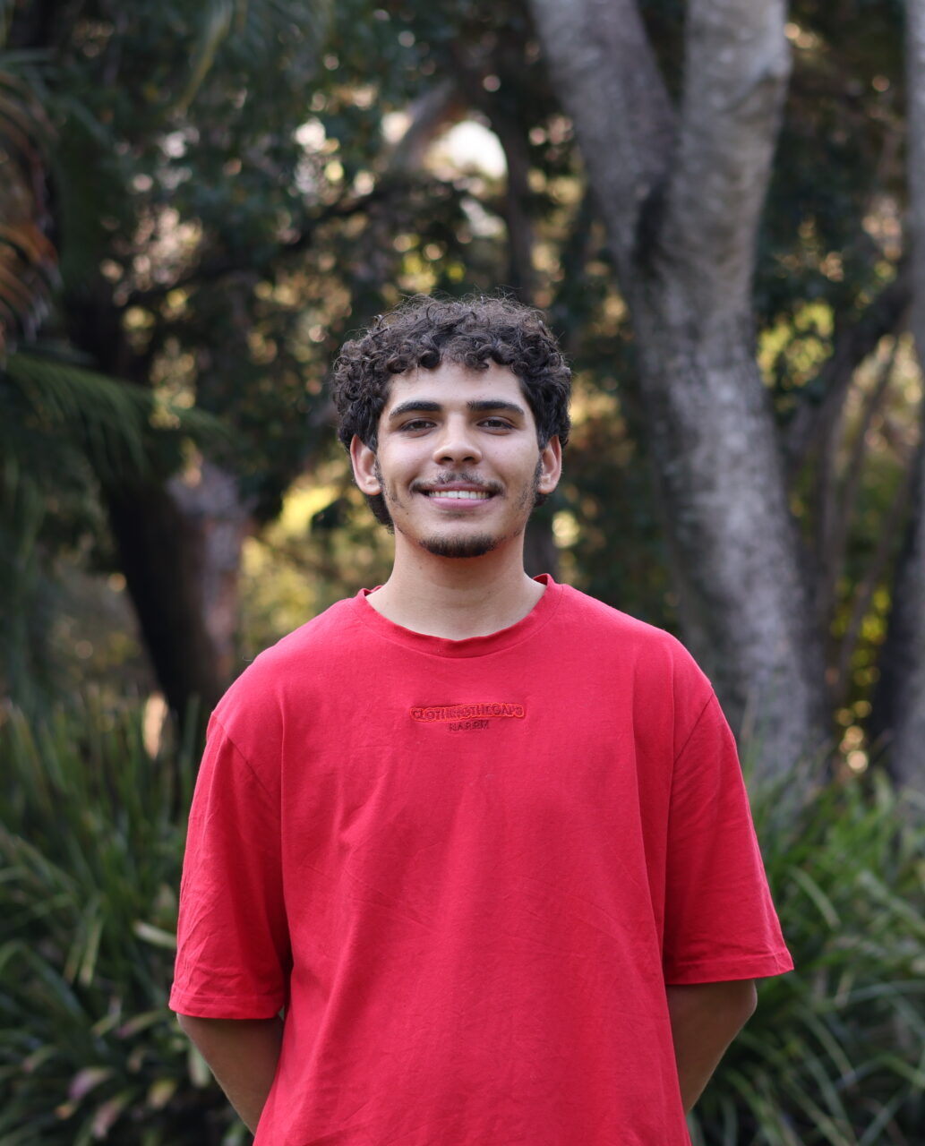 Kihi stands in natural light outside. They wear a red shirt, and are standing in front of trees.